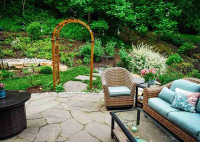 Cozy backyard patio with wicker furniture, cushions, and a stone floor, featuring a wooden garden arch and lush greenery in the background.