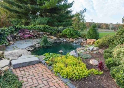 Beautiful backyard pond and garden design featuring a koi pond with a waterfall, stone patio with seating, brick walkway, and lush, vibrant landscaping.